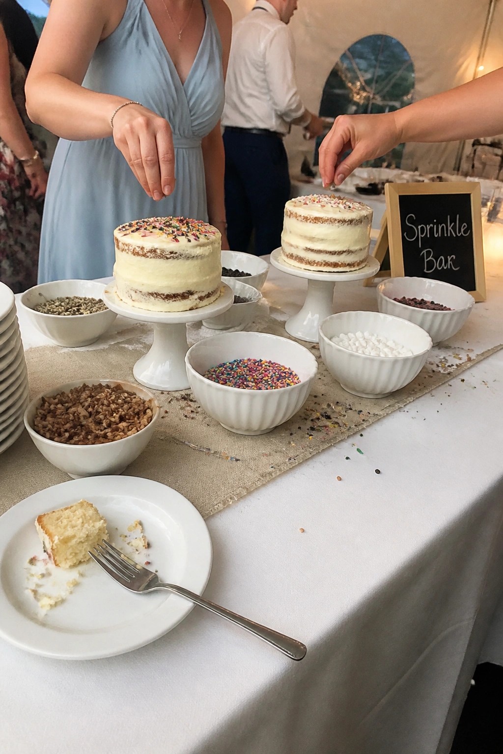 Two small cakes on pedestals surrounded by bowls of sprinkles and toppings at a wedding sprinkle bar with guests reaching in and a chalkboard sign visible.