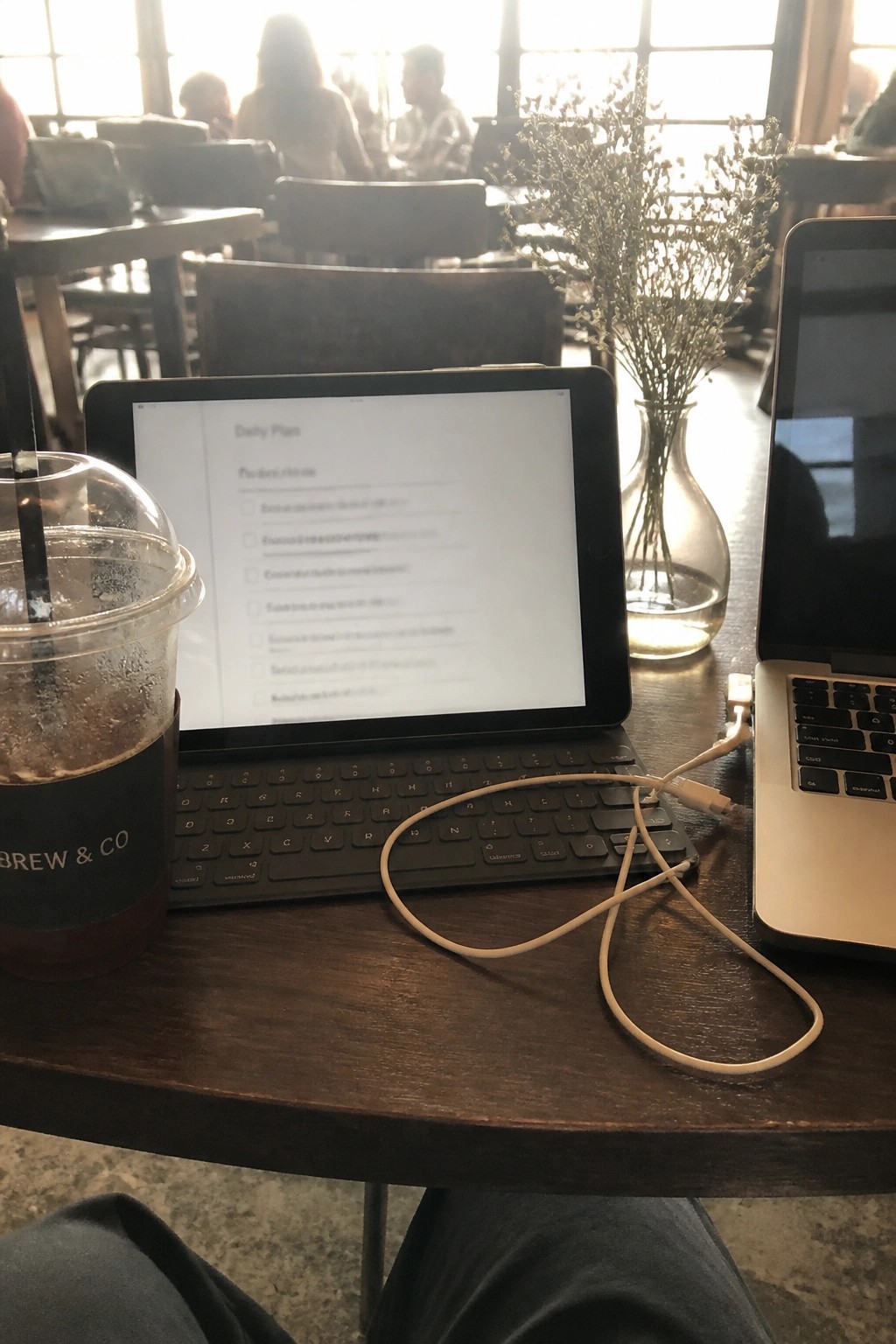 Wooden cafe table with tablet displaying daily plan checklist next to laptop and coffee cup in bright sunlight.