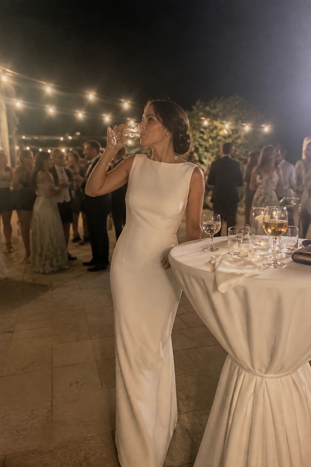 A woman in a minimalist white wedding dress drinks from a glass at a reception table under string lights with guests in the background.