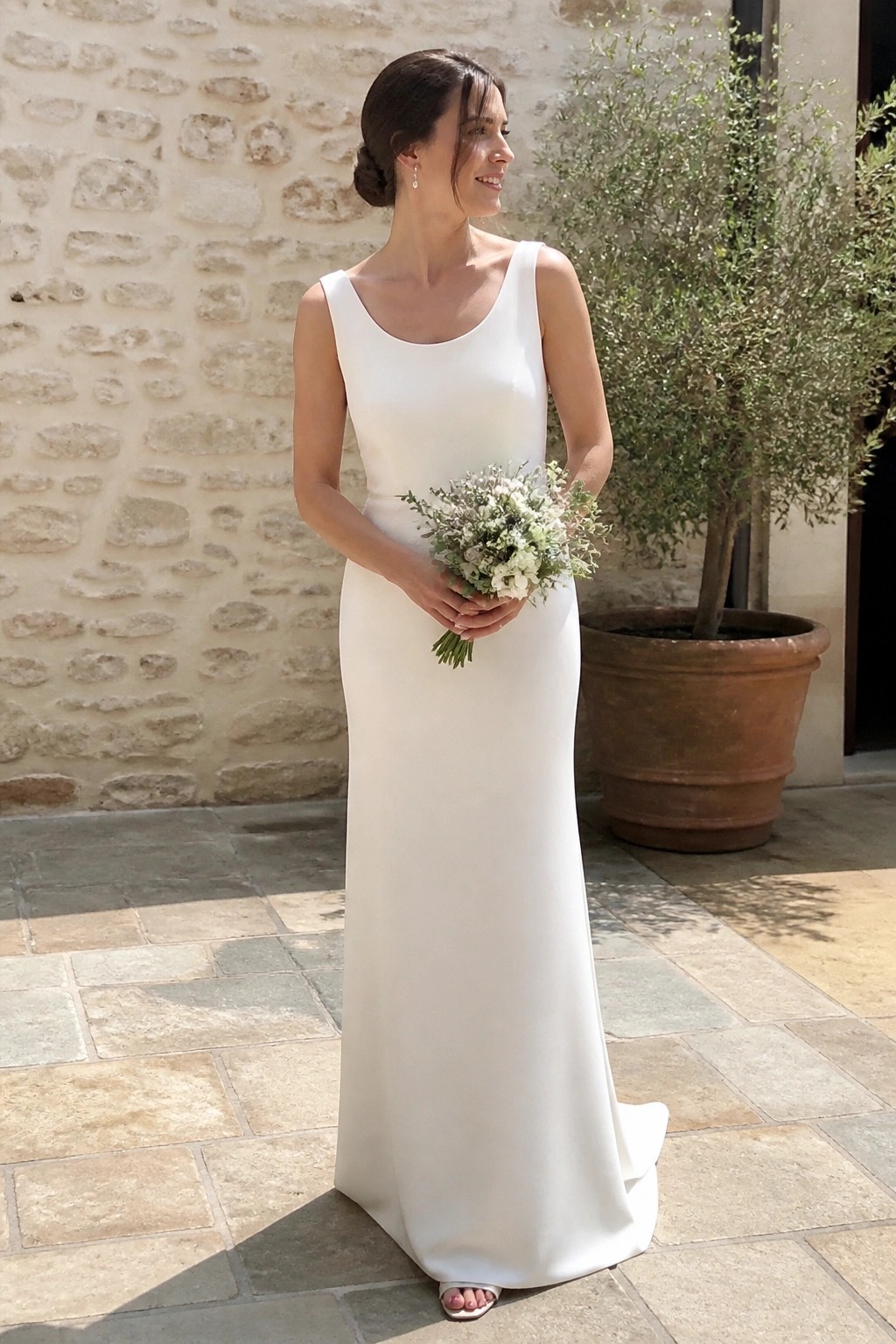 A bride in a minimalist white sheath wedding dress holds a wildflower bouquet on a stone patio with a rustic wall behind her.