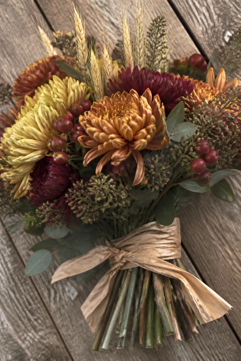 Close up of a rustic autumn wedding bouquet featuring yellow orange and burgundy chrysanthemums wheat stalks red berries and eucalyptus tied with a brown paper ribbon on a wooden surface.