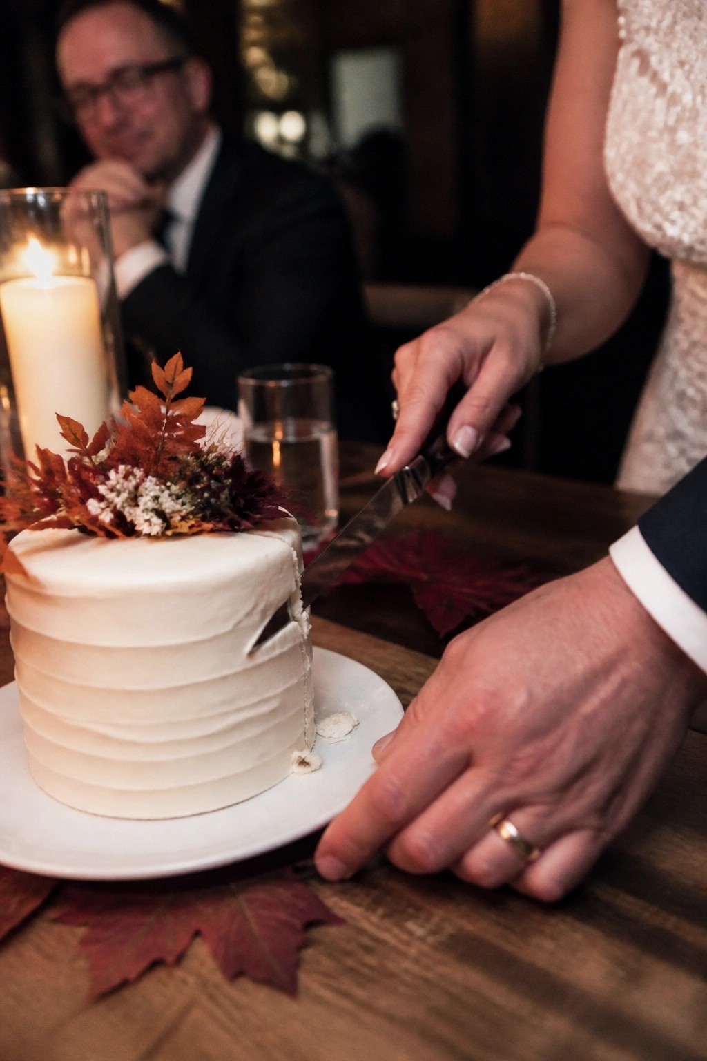 Bride and groom cutting a white wedding cake topped with autumn leaves during a candlelit rustic reception.