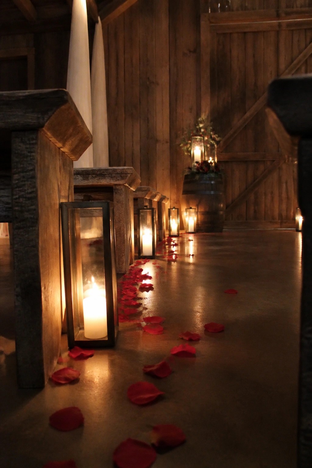 A dimly lit barn aisle lined with glowing lanterns and scattered red rose petals leading toward wooden doors and a floral barrel arrangement.