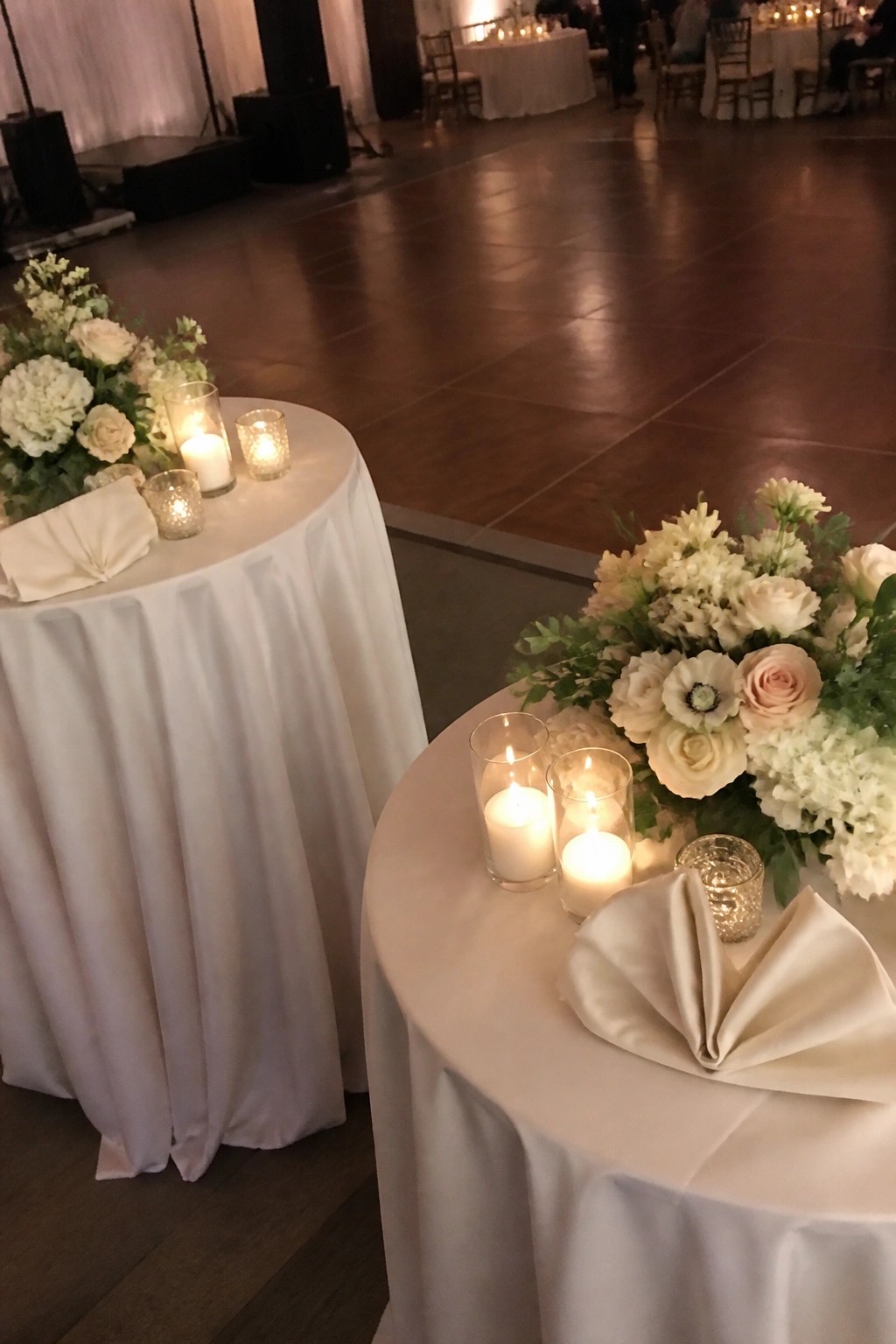 Two round tables with white tablecloths floral arrangements of white and blush flowers and lit candles in glass holders placed next to a wooden dance floor at a wedding reception.