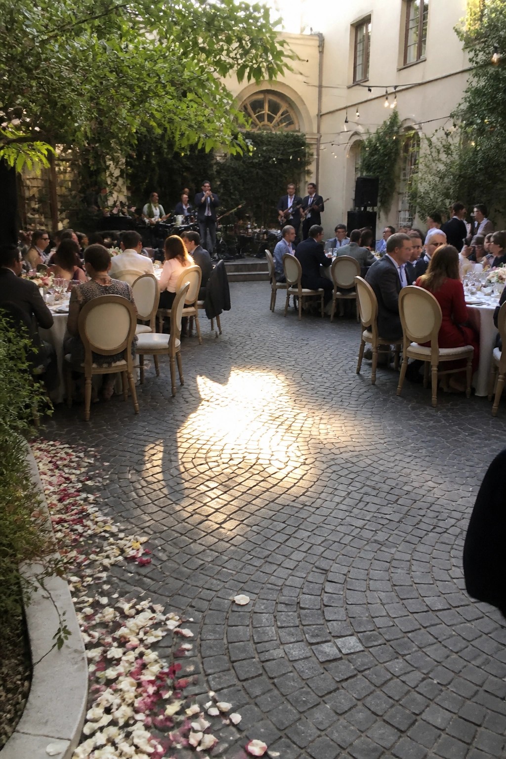 Elegant outdoor wedding reception in a sunlit historic courtyard with round guest tables, a live band on stage, string lights, and rose petals scattered on cobblestone.