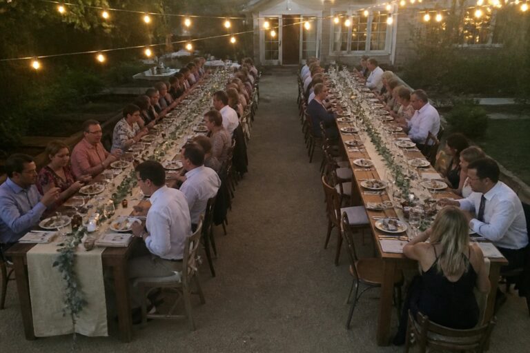 22 Dreamy Wedding Reception Ideas That Create Better Seating, Lighting, and Guest Flow Long wooden table set for an outdoor wedding reception with string lights overhead candles and seated guests at dusk