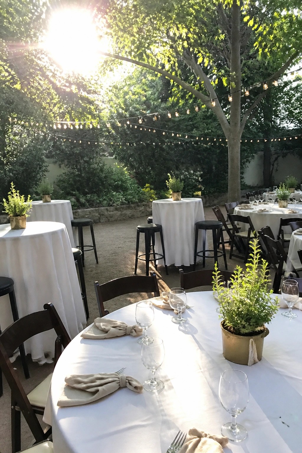 Outdoor wedding reception with white tablecloths round tables wooden chairs black stools potted green centerpieces and string lights hanging among trees in sunlight