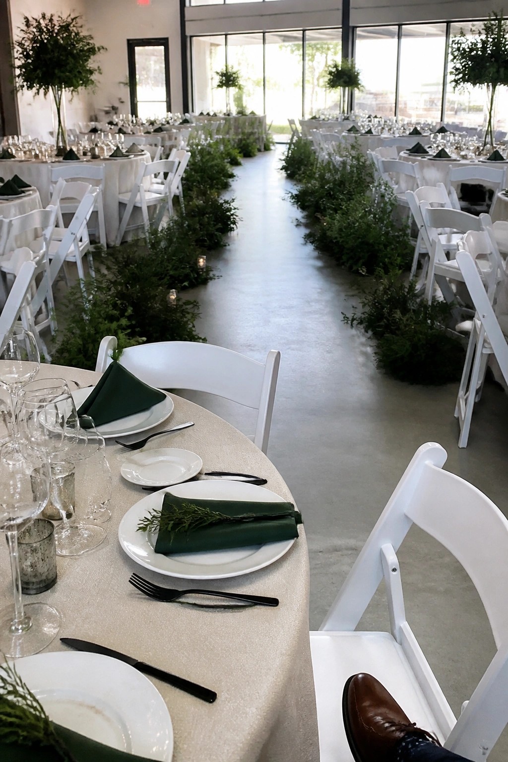 Aerial view of a wedding reception with white tables and chairs arranged around a central aisle lined with lush green plants and small lights in a bright hall with large windows