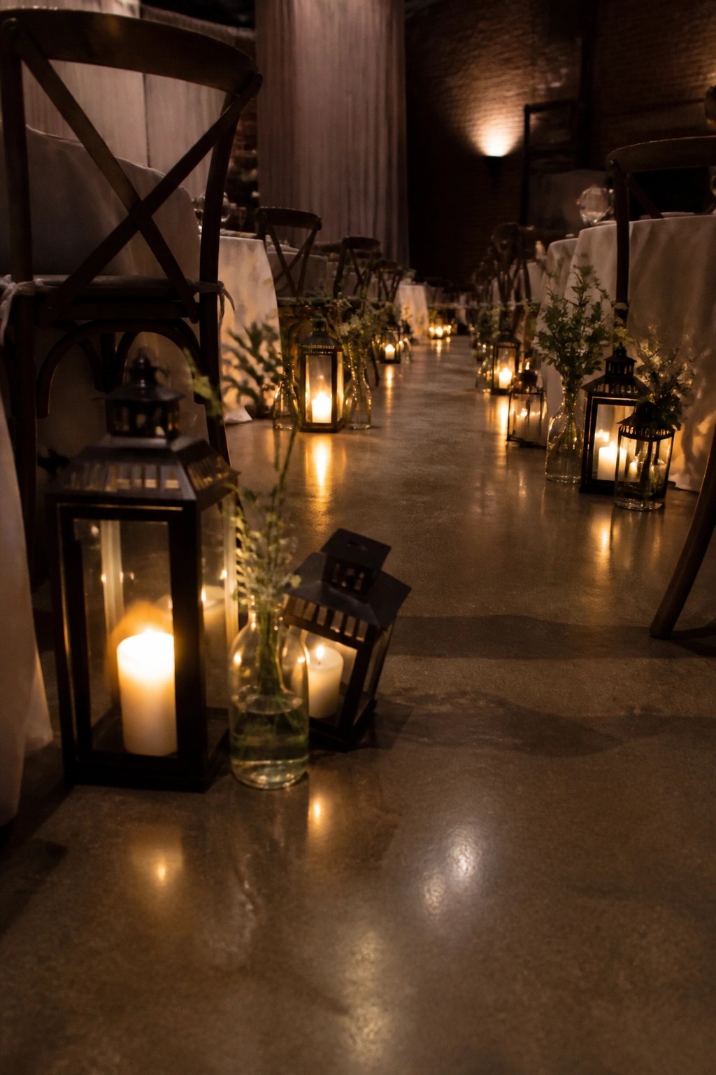 A romantic wedding reception aisle lined with glowing black lanterns and flower filled glass bottles on a concrete floor between white draped tables in a brick walled venue.