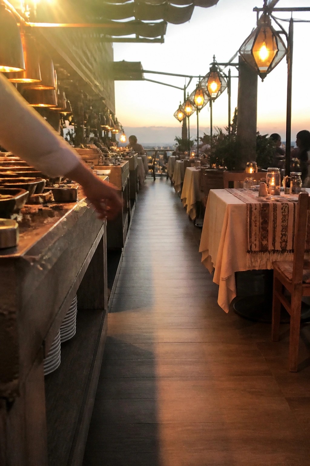 Outdoor wedding reception terrace at sunset with hanging lanterns, long buffet station on the left, and wooden tables set with white linens along a wide aisle leading to mountain views.