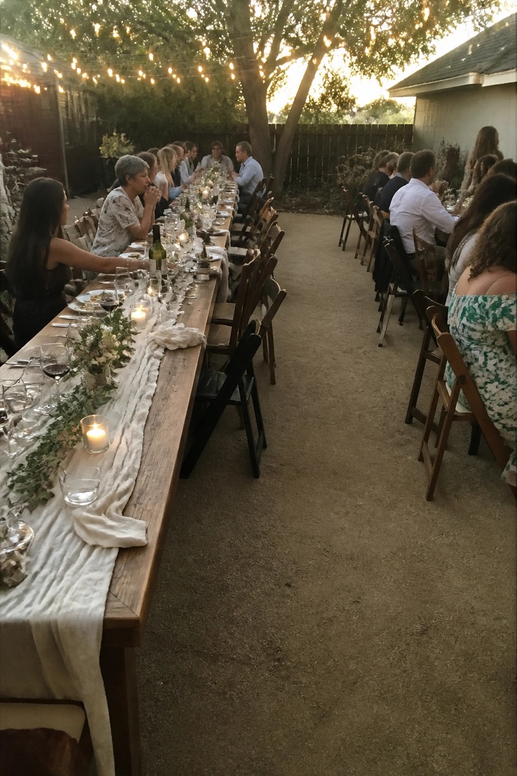 Long wooden table set for an outdoor wedding reception with string lights overhead candles and seated guests at dusk