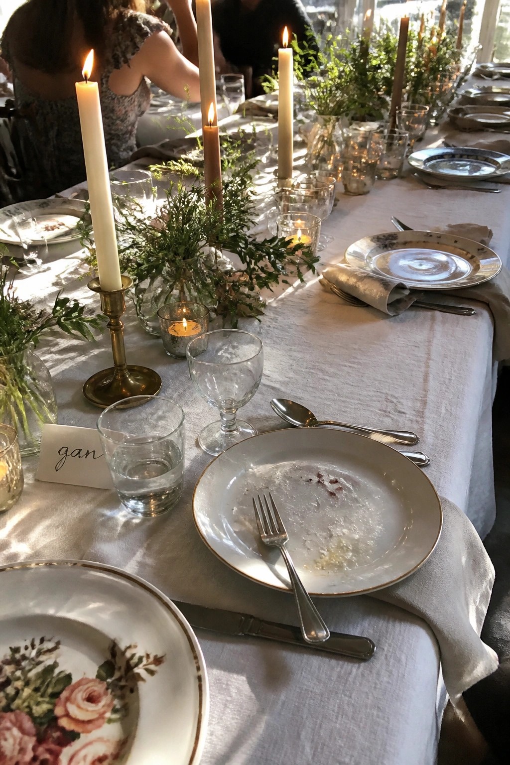 Long wedding table with white linens, lit taper candles in brass and glass, greenery centerpieces, floral plates, and natural window light.