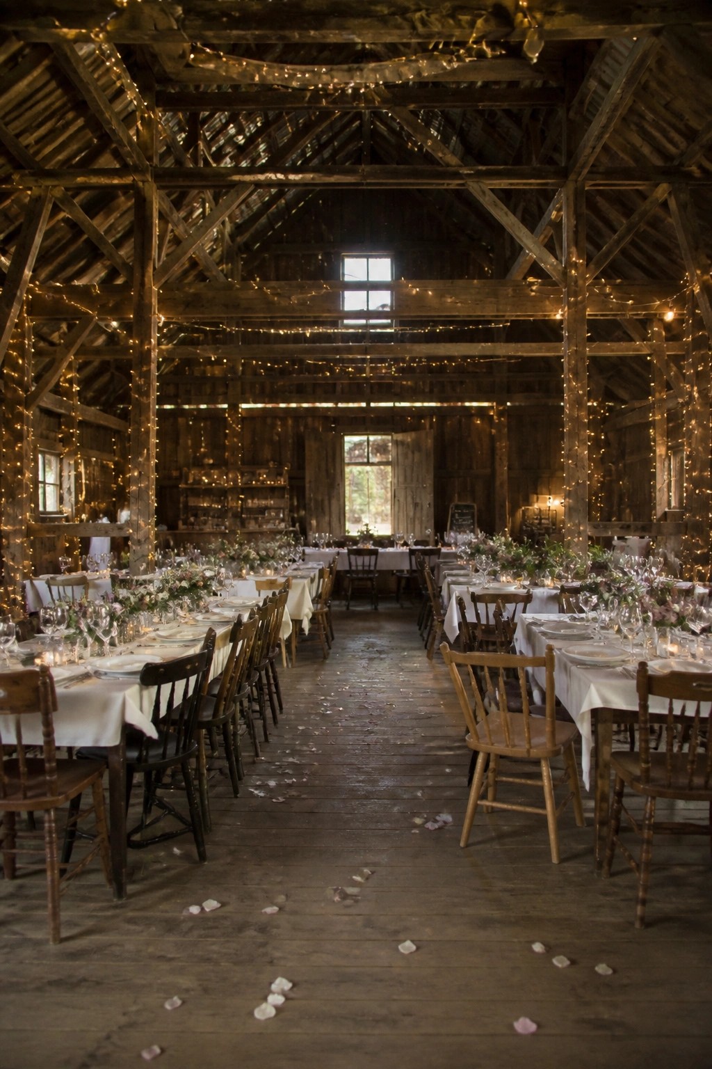 Interior of a rustic wooden barn wedding reception with long white tablecloths, mixed wooden chairs, hanging string lights, floral arrangements, and rose petals on the floor at dusk.