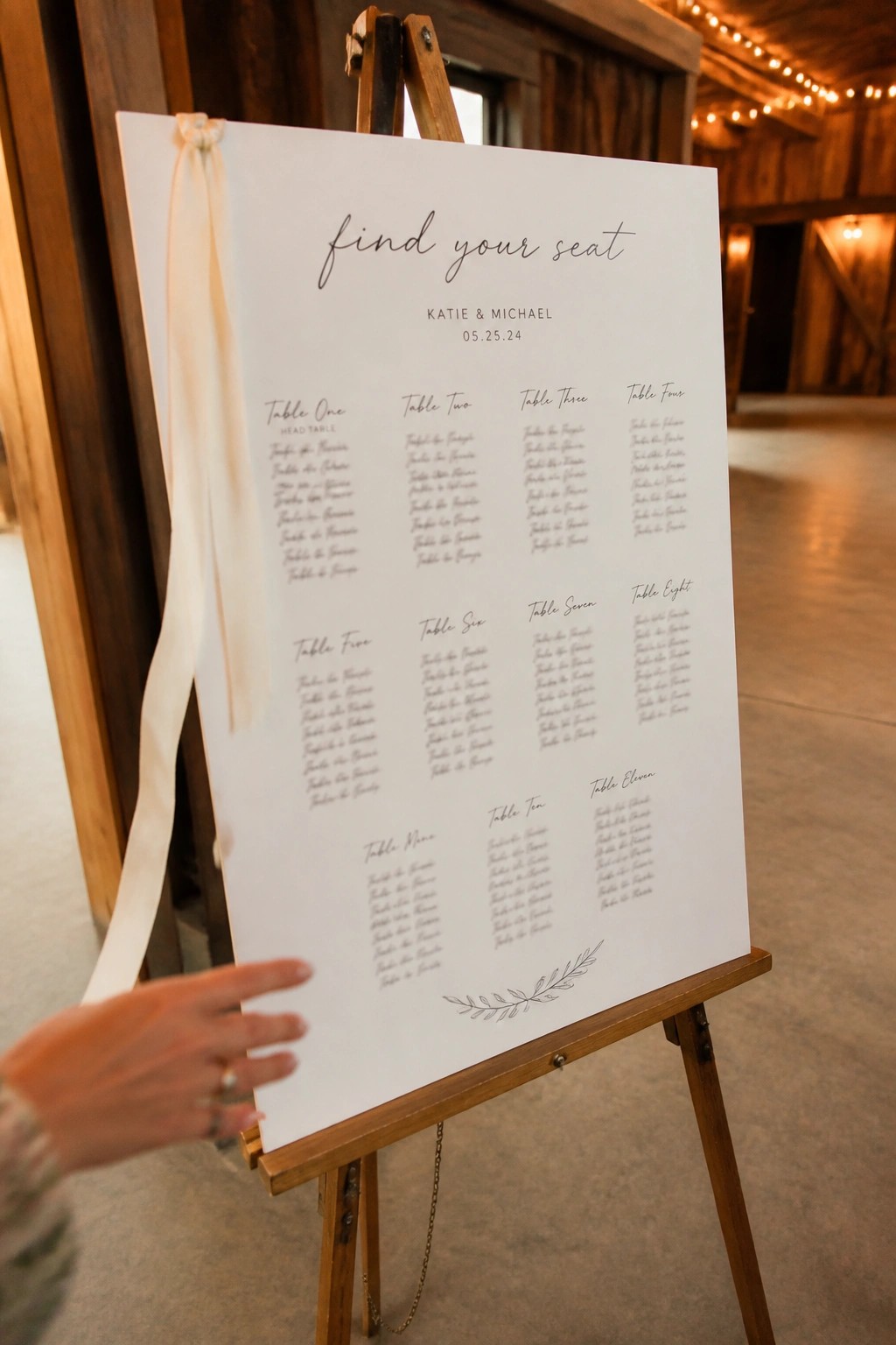 A white seating chart sign on a wooden easel in a rustic barn wedding venue with string lights and a hand pointing to table assignments for Katie and Michael.