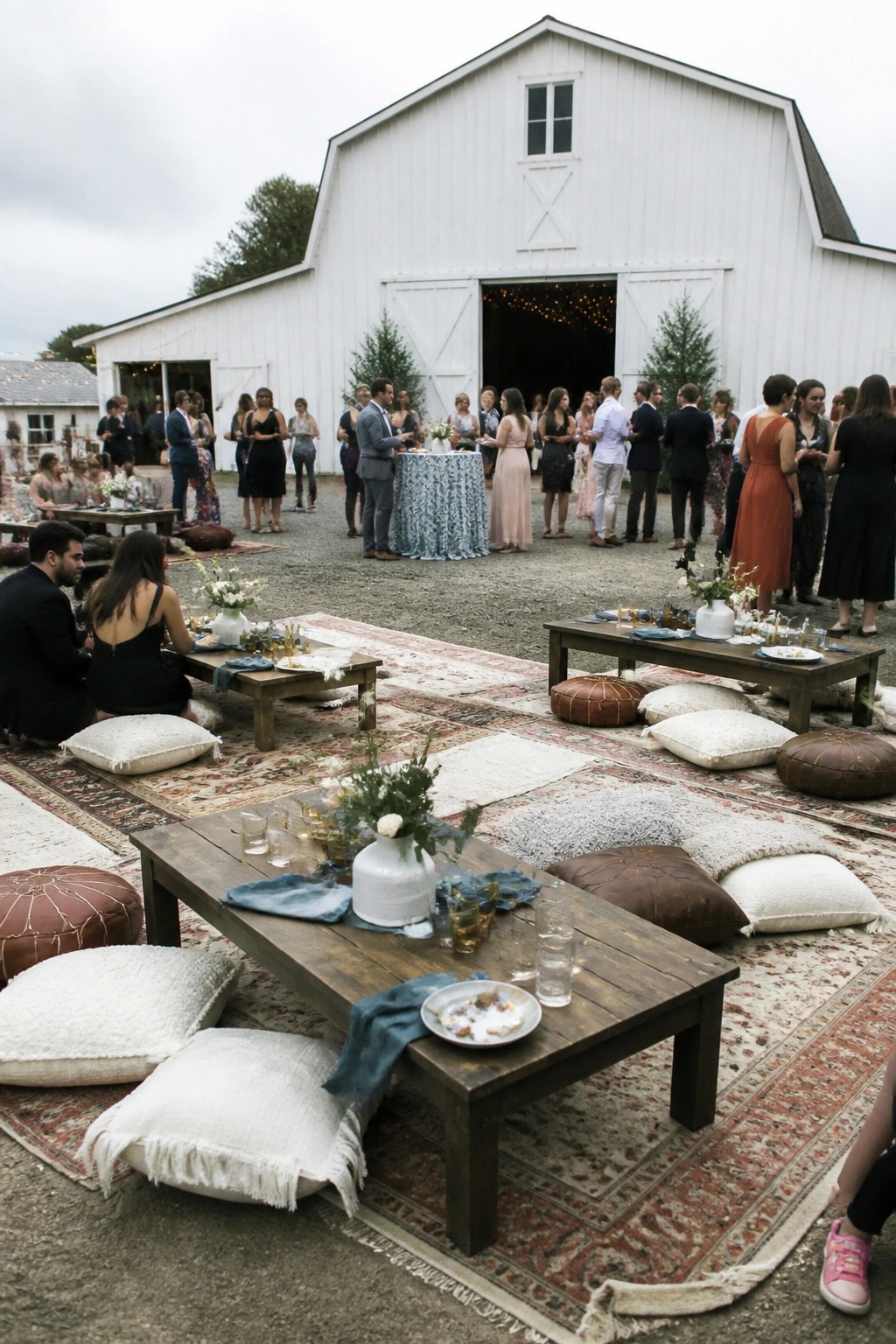 Wedding reception with low wooden tables and layered rugs holding pillows and poufs outside a white barn with guests mingling nearby