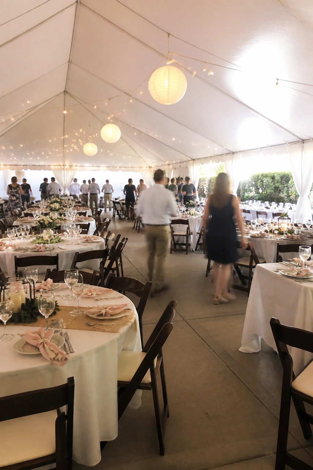 Large white tent wedding reception with round tables, pink napkins, floral centerpieces, hanging paper lanterns, and string lights as guests mingle in the open space.