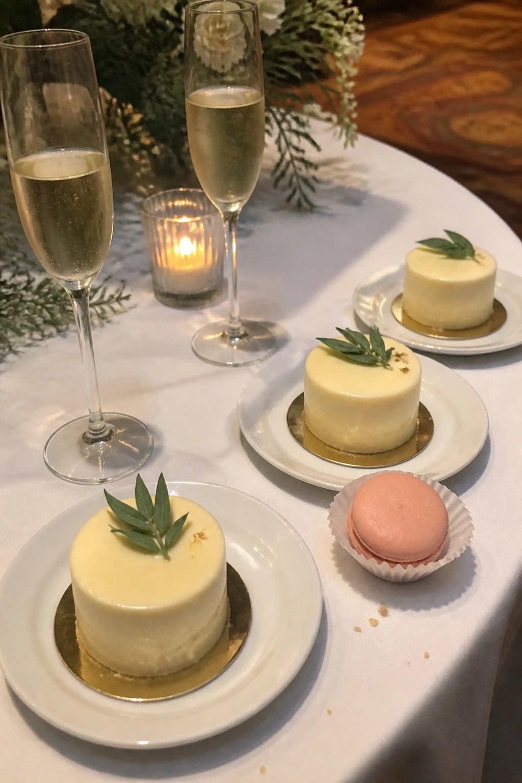 Elegant wedding table with two champagne flutes a lit candle three ivory mini cakes topped with sage and one blush macaron on white plates with gold accents and floral greenery in the background.