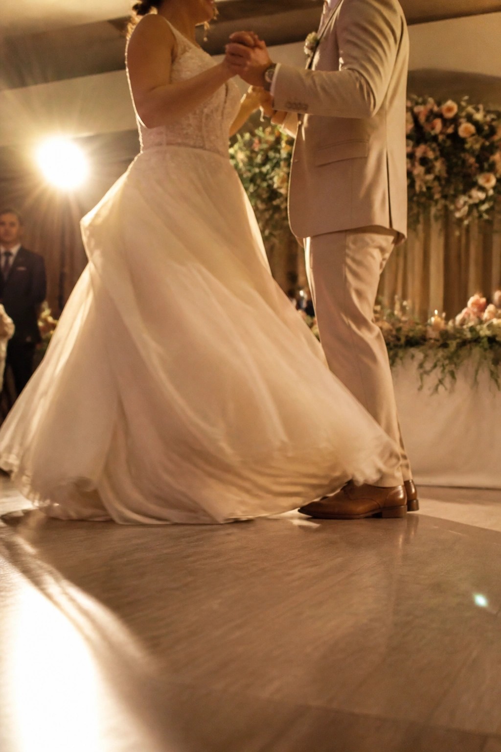 Bride in flowing ivory wedding dress and groom in champagne suit share a first dance on a polished floor with soft blush and sage floral arrangements in the background.