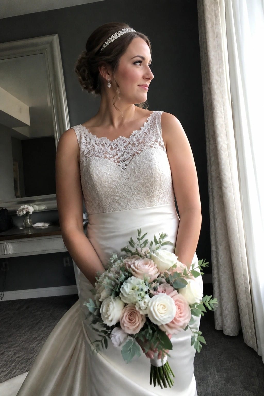 A bride in an ivory lace and satin wedding gown holds a bouquet of white and blush roses with sage greenery while standing in a gray walled room.