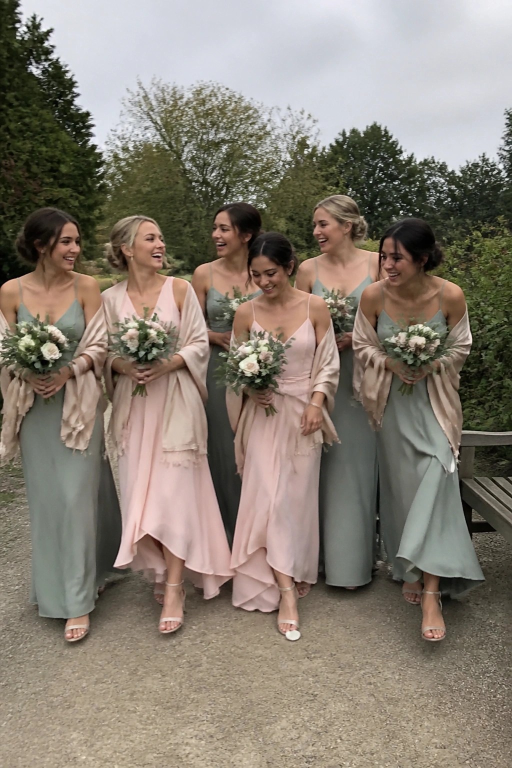 Bridesmaids in sage green and blush pink dresses with champagne wraps holding floral bouquets in a garden setting