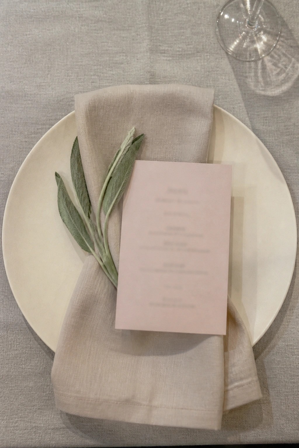 A wedding place setting featuring an ivory plate topped with a folded champagne napkin, sage green leaves, and a soft blush menu card on a neutral linen tablecloth.
