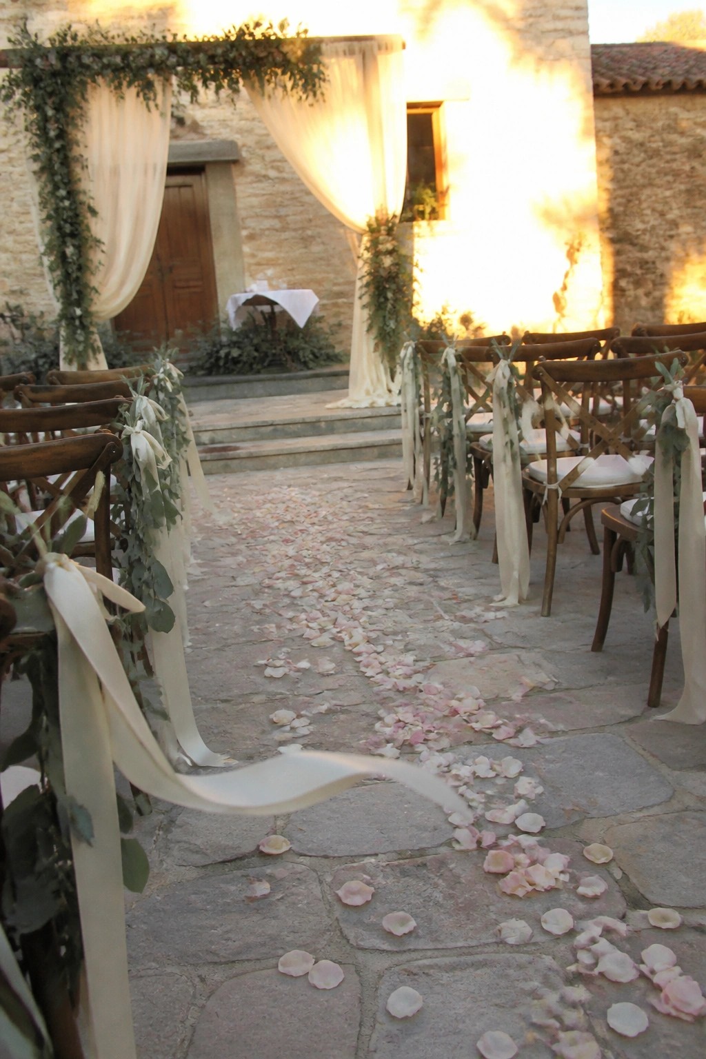 Stone courtyard wedding aisle lined with wooden chairs tied in ivory ribbons and greenery with blush rose petals scattered on the path leading to a draped arch at sunset
