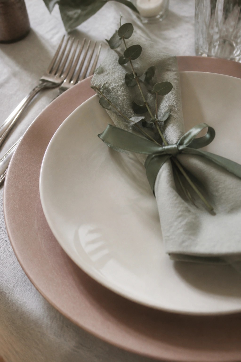 Close up of blush pink and white stacked plates with a sage green napkin tied by a ribbon and eucalyptus on a white tablecloth in soft natural light.