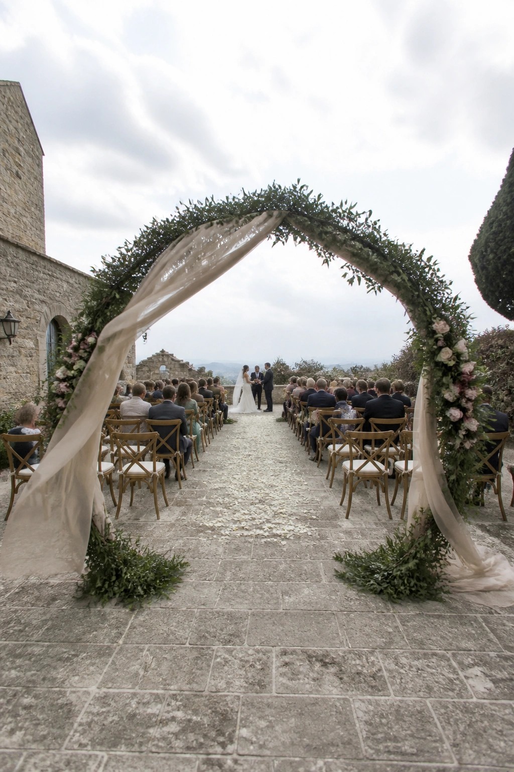 Outdoor wedding ceremony on a stone terrace with a large arch of greenery and pink roses, white drapes, wooden chairs, and guests facing the couple against a scenic landscape.