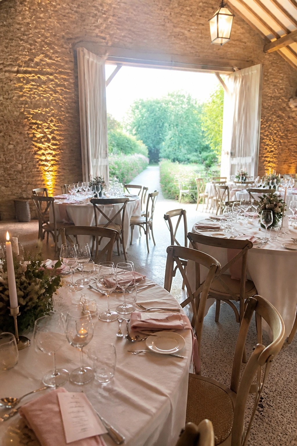 Elegant wedding tables in a rustic stone hall with open doors to a green garden, featuring white linens, pink napkins, wooden chairs, candles, and floral centerpieces.