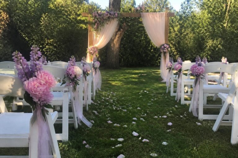 Wooden wedding arch with white draping and pastel pink purple flowers beside decorated white chairs on a grassy lawn scattered with petals.