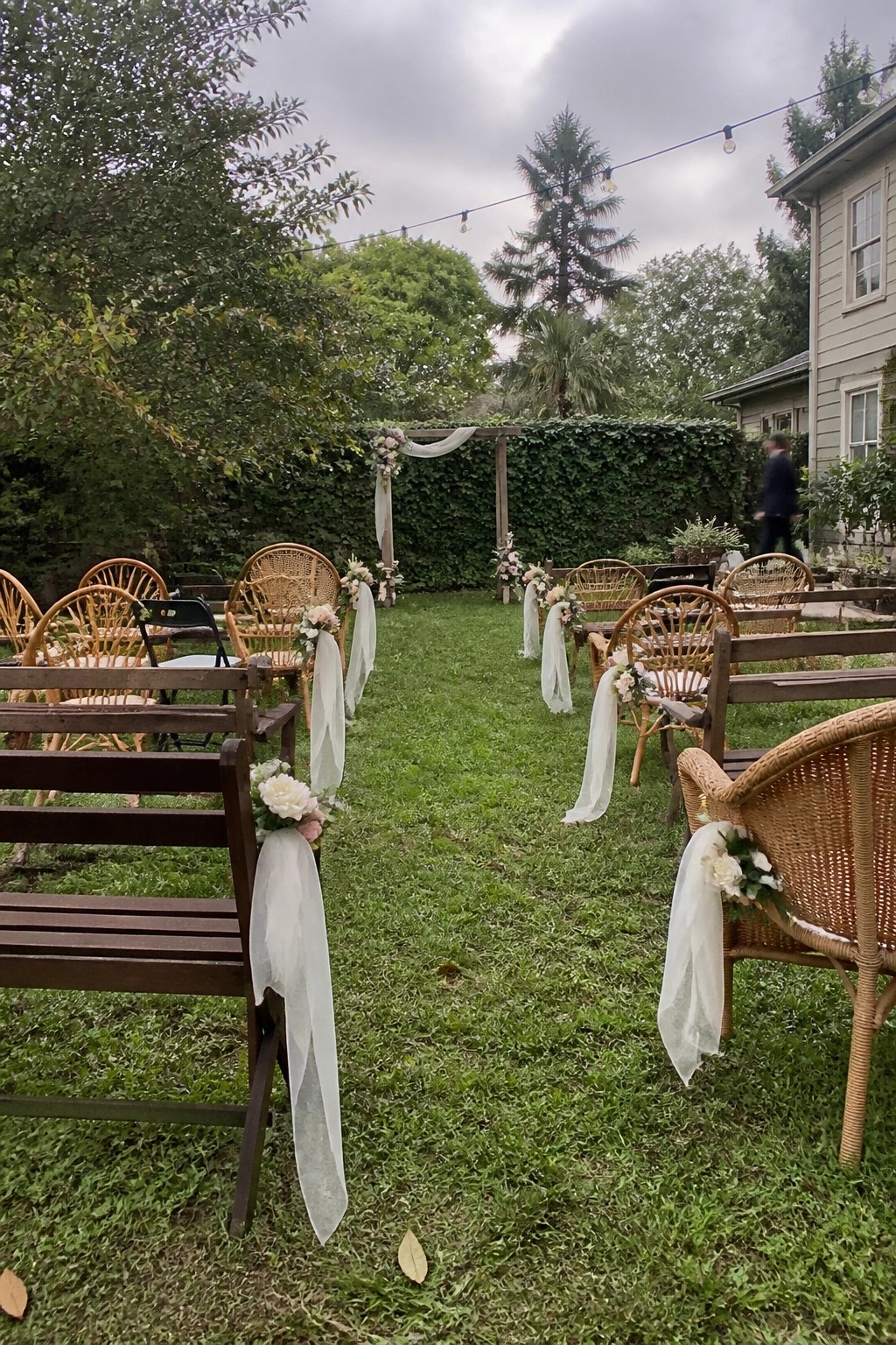 Outdoor wedding aisle lined with decorated wicker chairs and wooden benches leading to a floral arch in a lush garden setting.