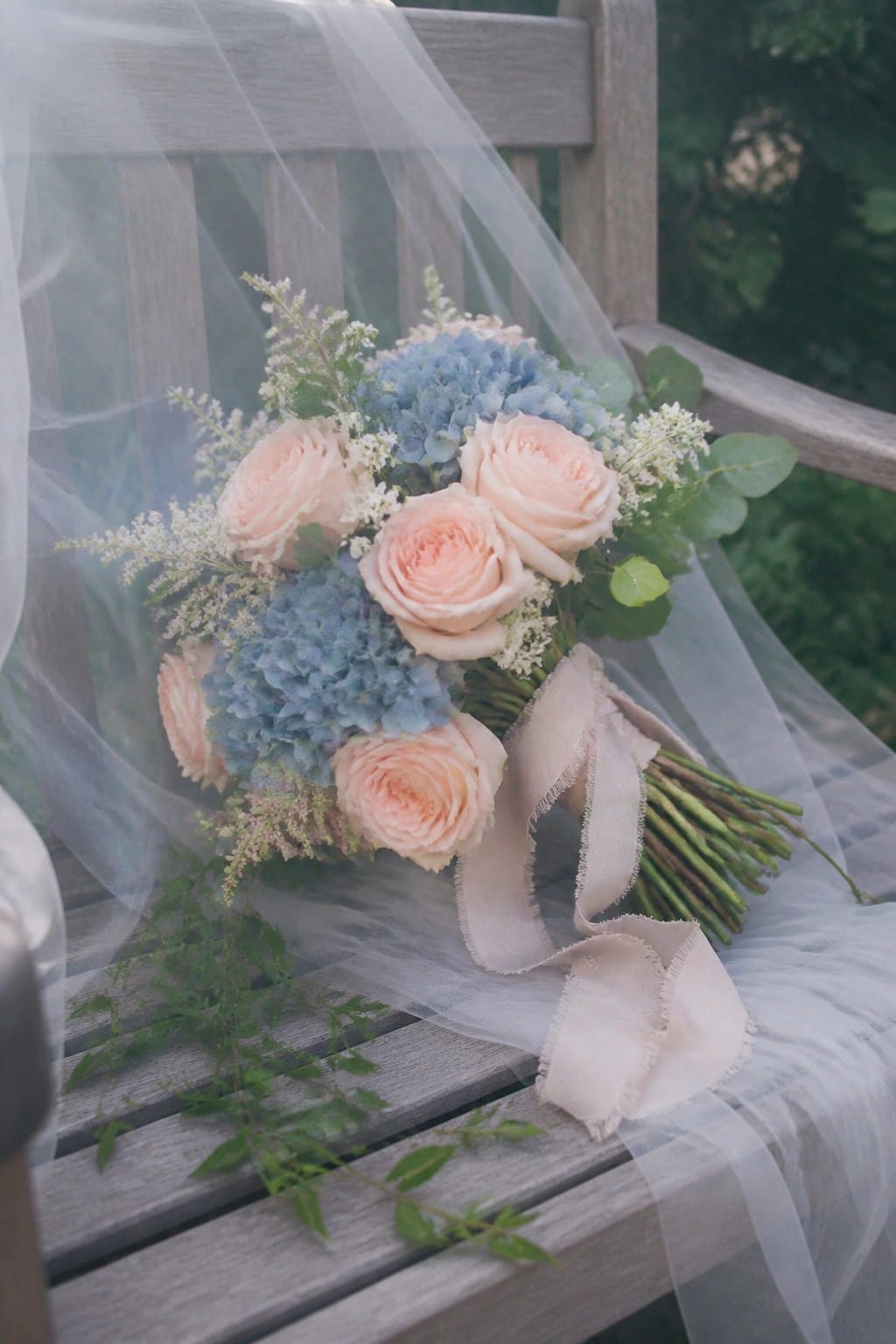 A bouquet of peach roses and blue hydrangeas with sheer white tulle draped over a wooden bench in a garden setting.