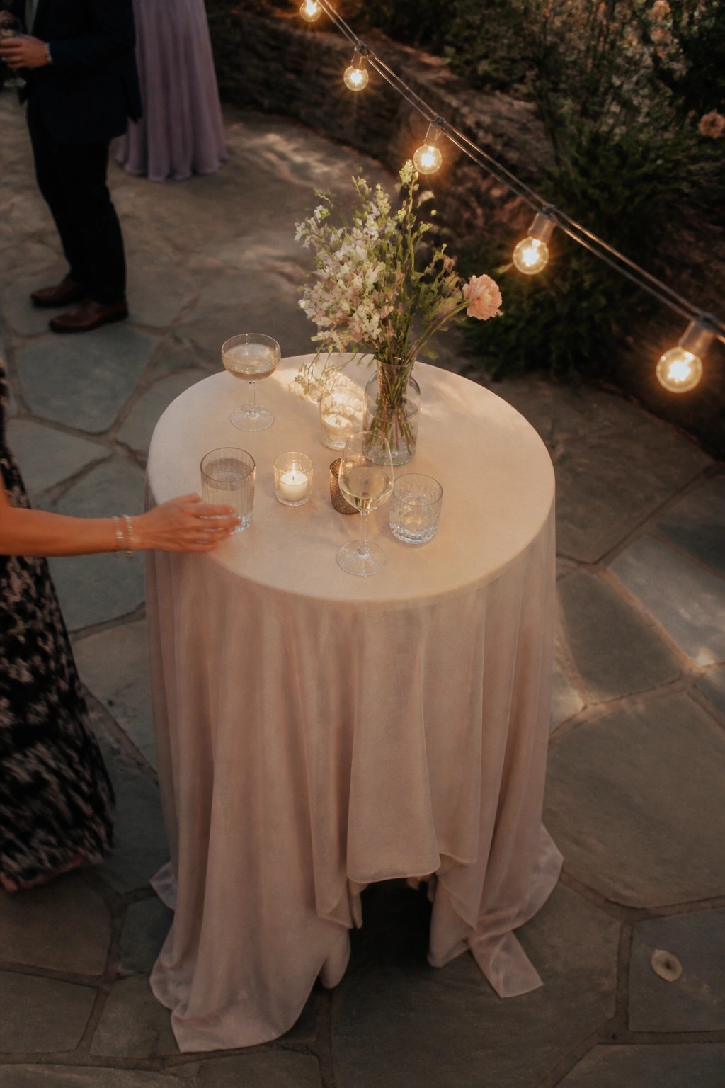 Round table with blush draping, pink and white floral centerpiece, candles, and glassware on a stone patio under string lights at dusk.