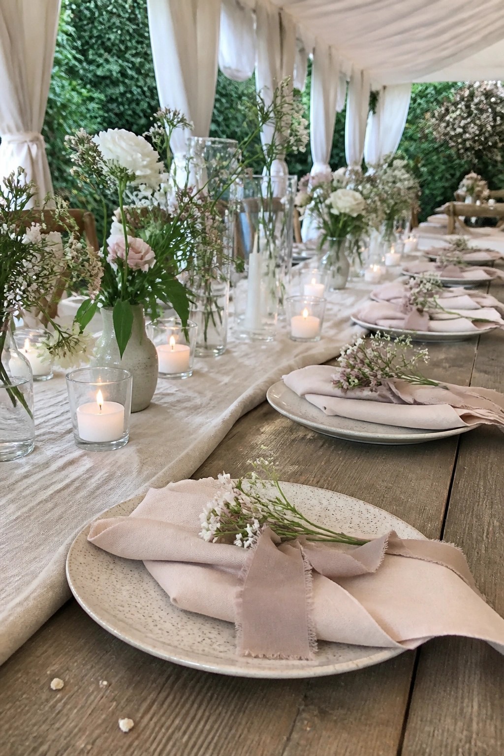 Long wooden wedding table with blush napkins on speckled plates, white and pink floral centerpieces, and glowing candles beneath white drapes in a garden setting.