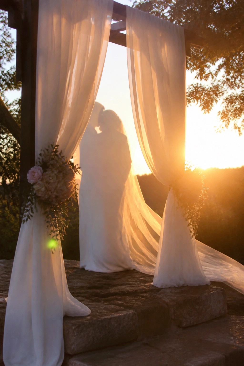 Silhouetted bride and groom kissing under flowing white drapes on a wooden arch decorated with pastel florals at golden hour sunset on stone steps outdoors.