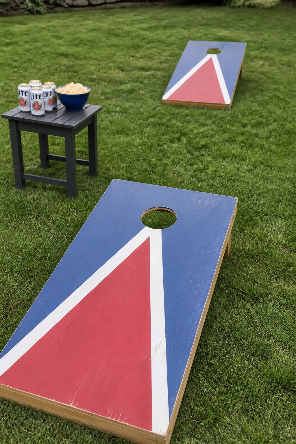 Two cornhole boards on a green lawn beside a table with beer cans and chips for wedding guests