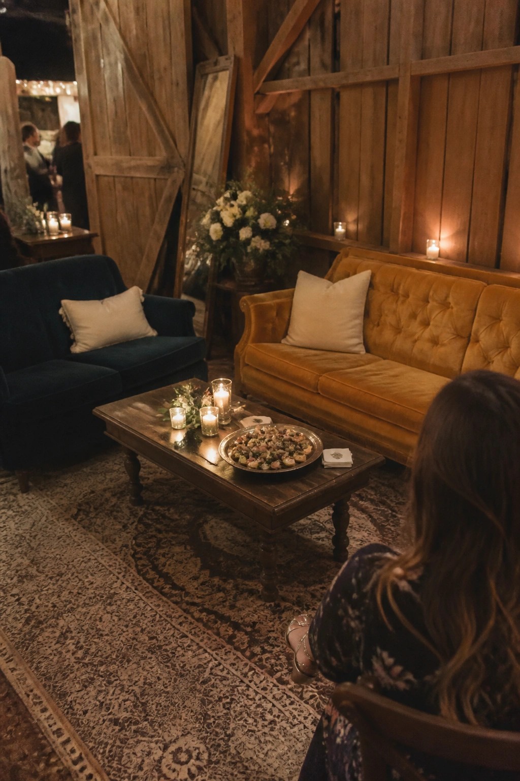 Cozy wedding lounge with blue and mustard velvet sofas, candlelit coffee table with appetizers, and rustic barn wood walls at night.