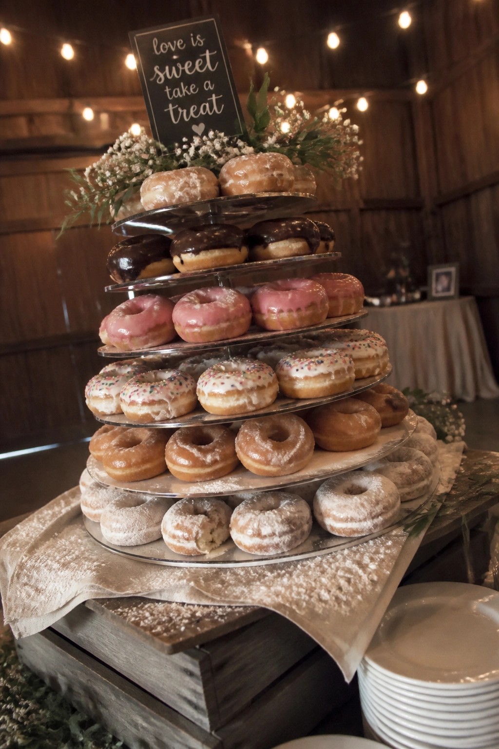 Multi tiered donut stand with assorted glazes and sprinkles in a rustic barn setting with string lights and a love is sweet sign