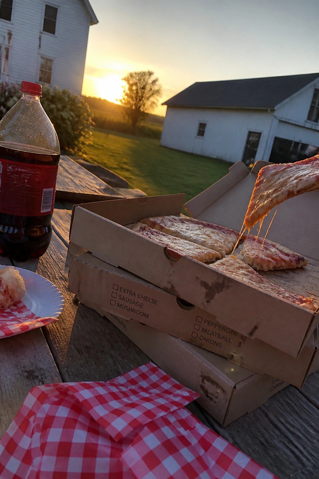 Open pizza boxes with stretching cheese on a wooden table at golden hour sunset with soda and checkered napkins on a farm lawn.