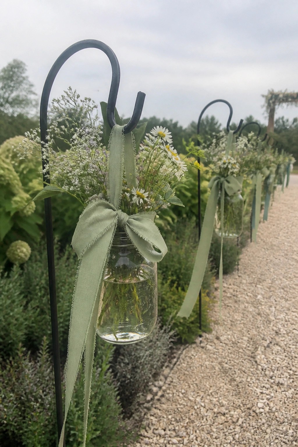 A row of mason jars filled with wildflowers and tied with sage green ribbons hanging from black hooks along a garden wedding path.