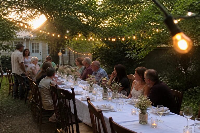 Long outdoor wedding table set with white linens, plates, and wildflowers in mason jars under glowing string lights in a backyard at dusk.