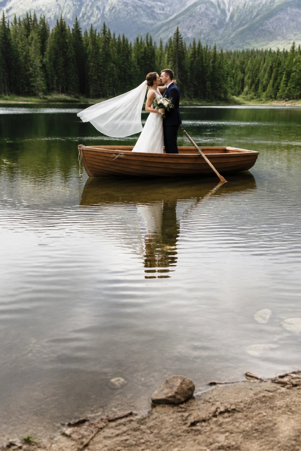 Bride and groom kissing in a wooden boat on a calm lake with mountains and forest behind them their reflection visible in the water
