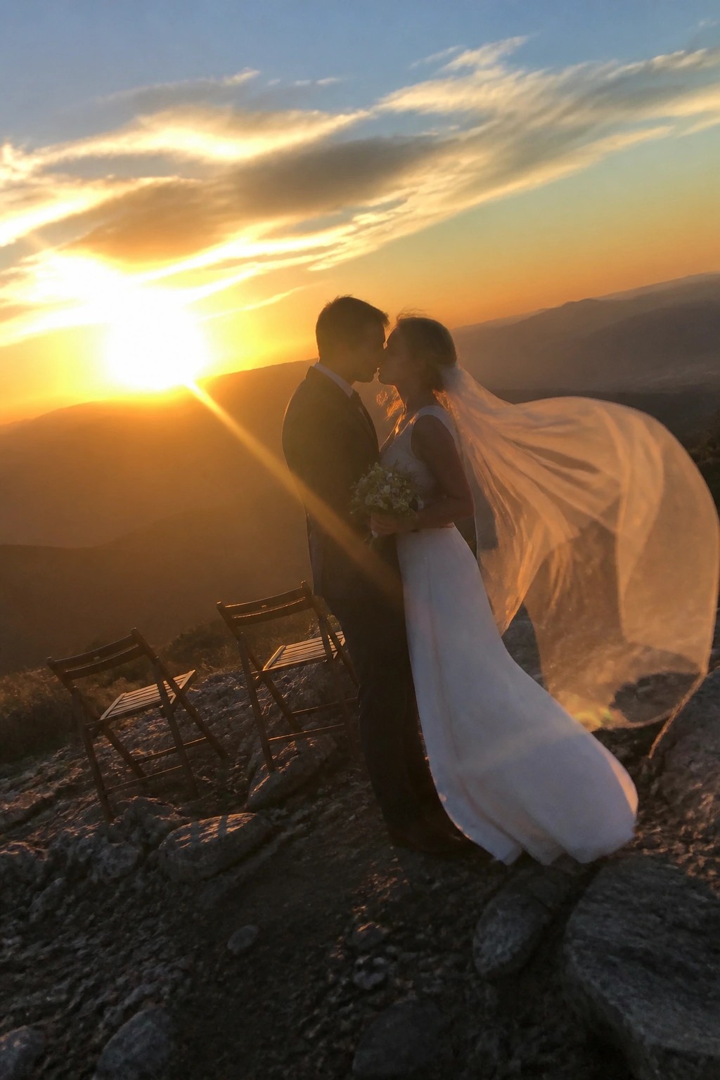 A silhouetted bride and groom kiss at sunset on a rocky mountain peak with the bride's veil flowing in the wind and two chairs nearby