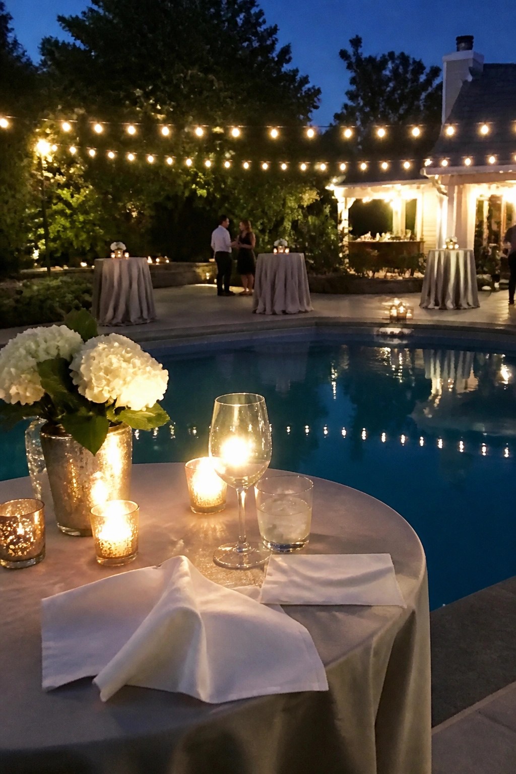 Evening wedding reception by a swimming pool with string lights candles and white floral arrangements on tables
