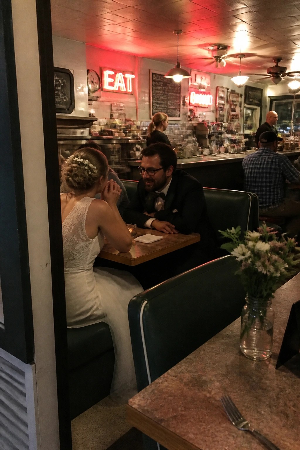 A bride in a lace wedding dress and groom in a suit share a booth in a retro diner lit by red neon signs at night.