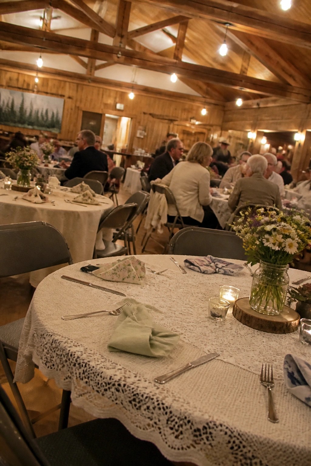 Rustic wedding reception inside a wooden barn hall with round tables covered in lace cloths, wildflower mason jar centerpieces, and guests seated under warm overhead lights.