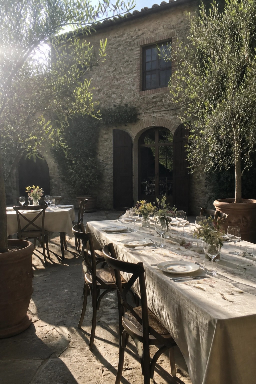 Long wedding table set with white linens and flowers in a rustic stone courtyard surrounded by olive trees and wooden chairs under bright sunlight.