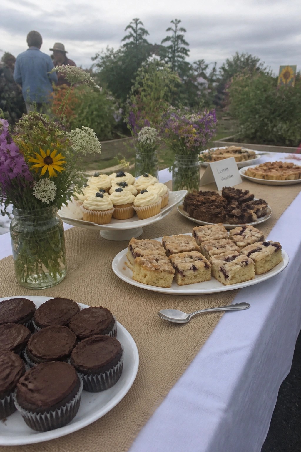 Outdoor wedding dessert table with assorted cupcakes cakes and wildflower mason jars in a garden setting
