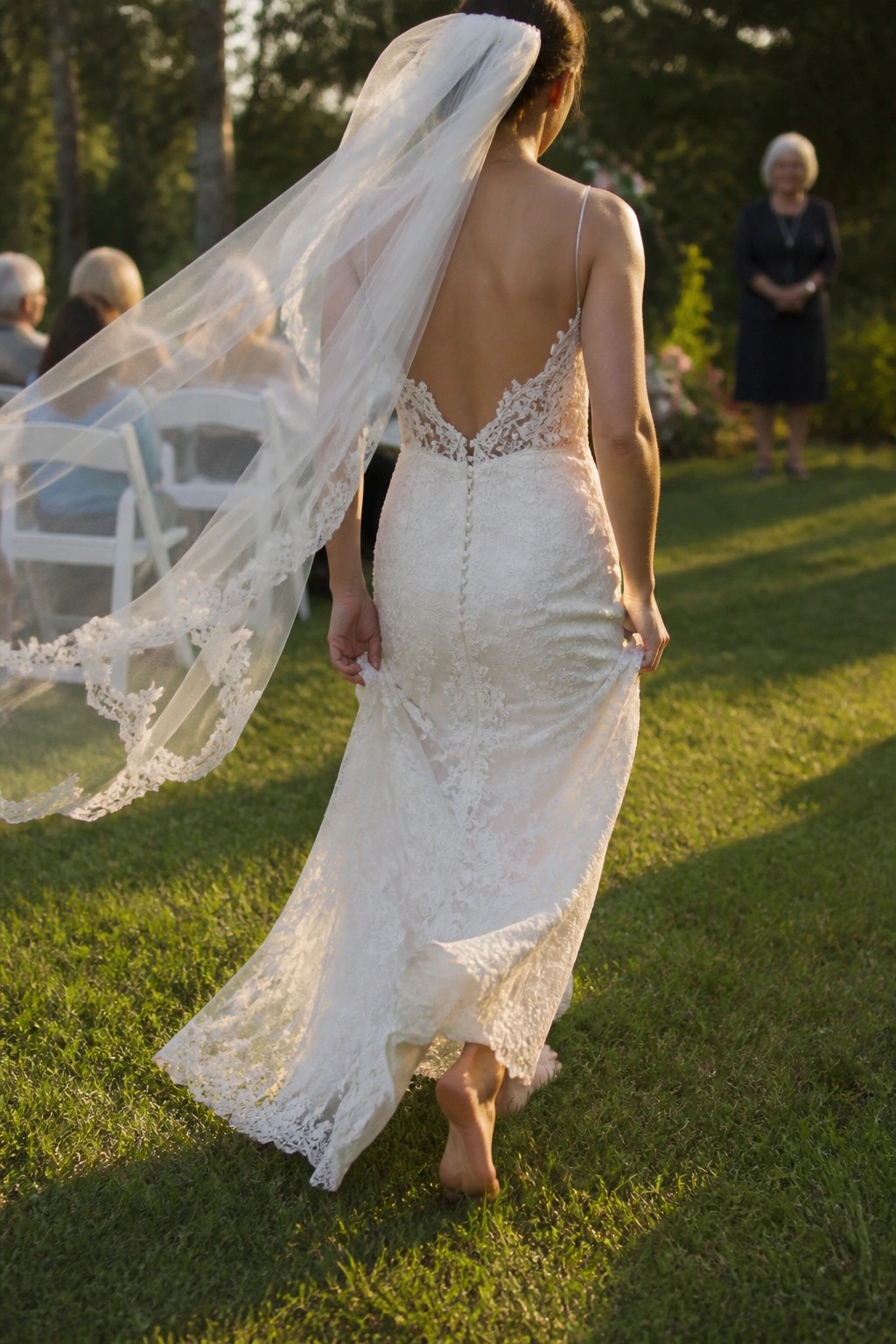Bride in lace gown with flowing veil walks barefoot on grass during backyard wedding at sunset with guests in background.