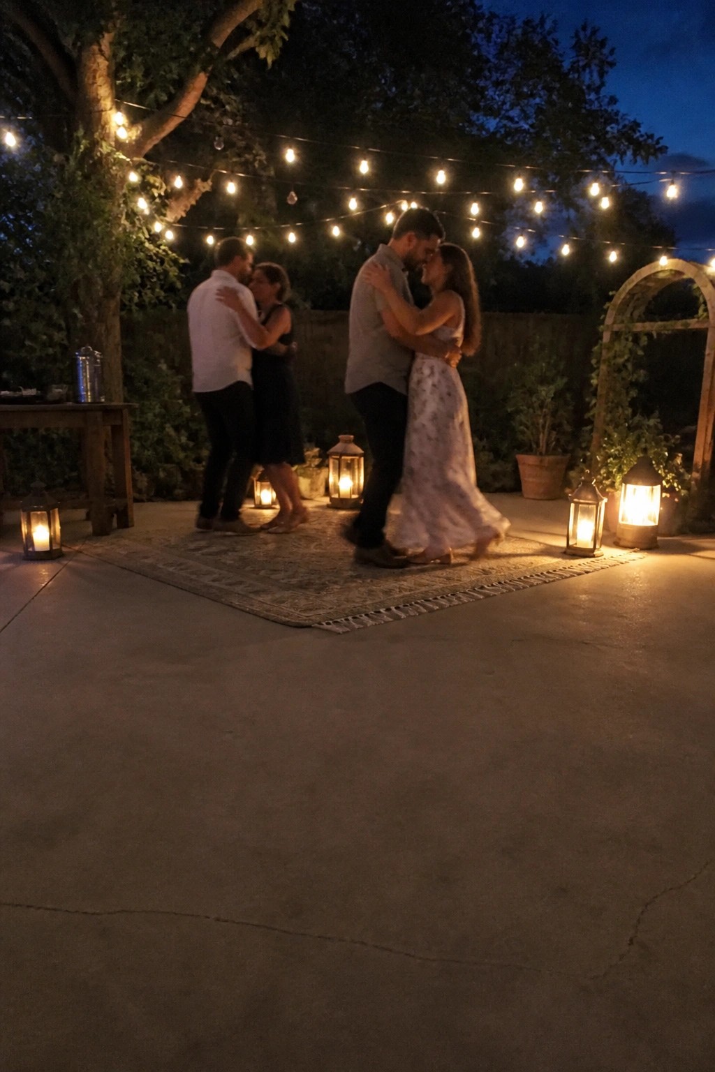Two couples slow dancing on a rug in a backyard at dusk with string lights and lanterns creating a romantic glow